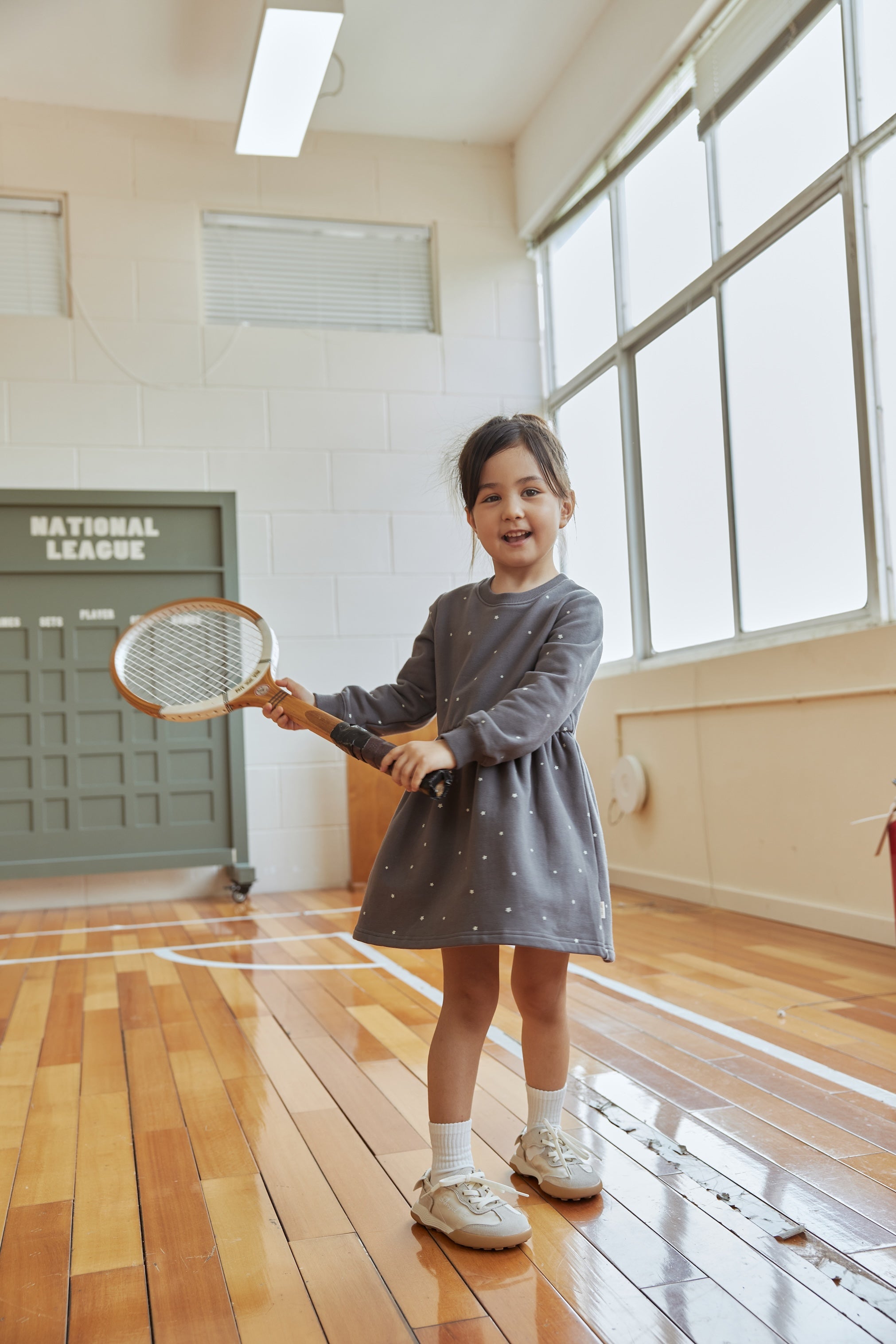 Young girl in a gray dress holding a tennis racket indoors.