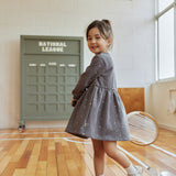 Young girl in a gray dress standing on a wooden floor with a green door labeled 'National League' in the background.