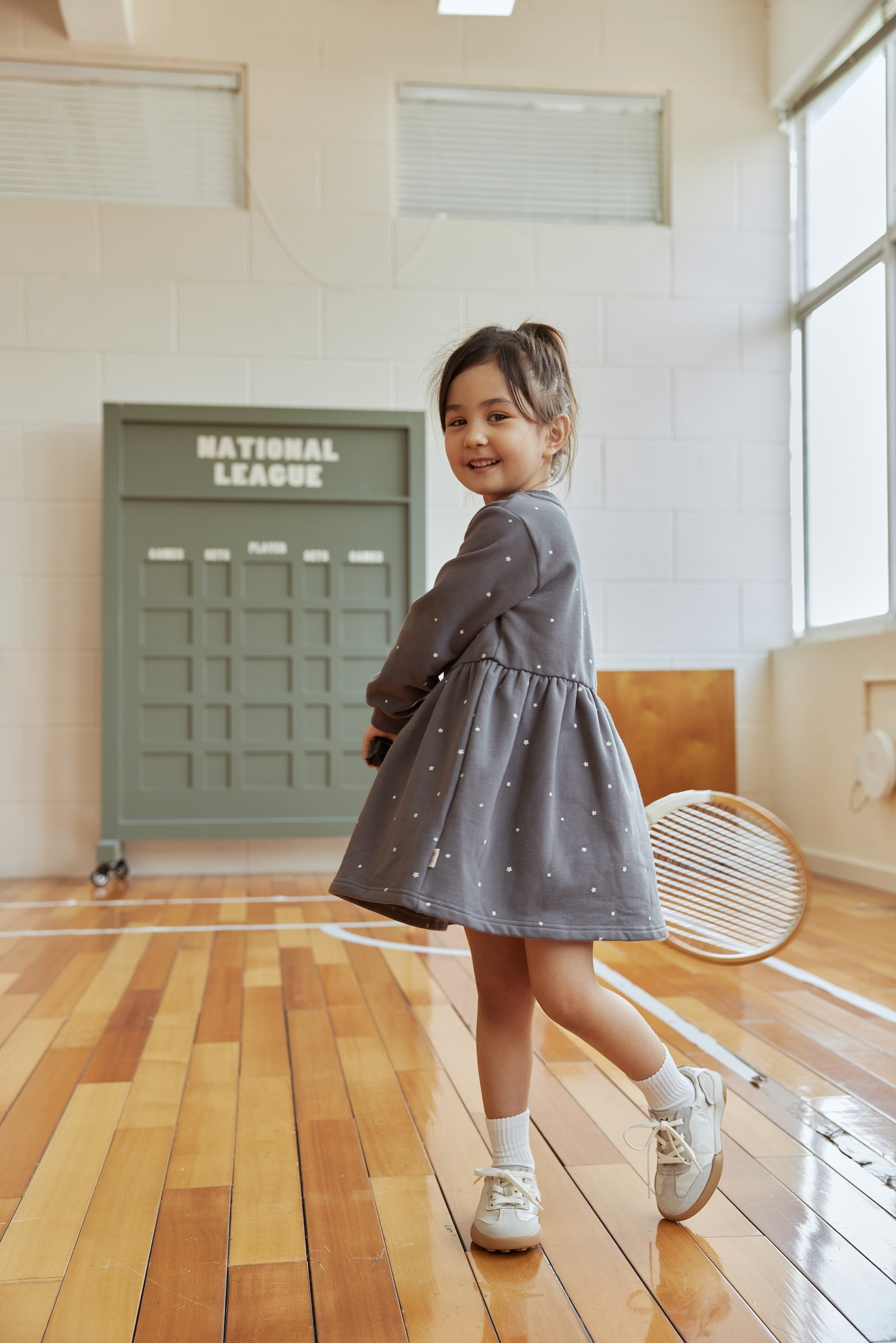Young girl in a gray dress standing on a wooden floor with a green door labeled 'National League' in the background.