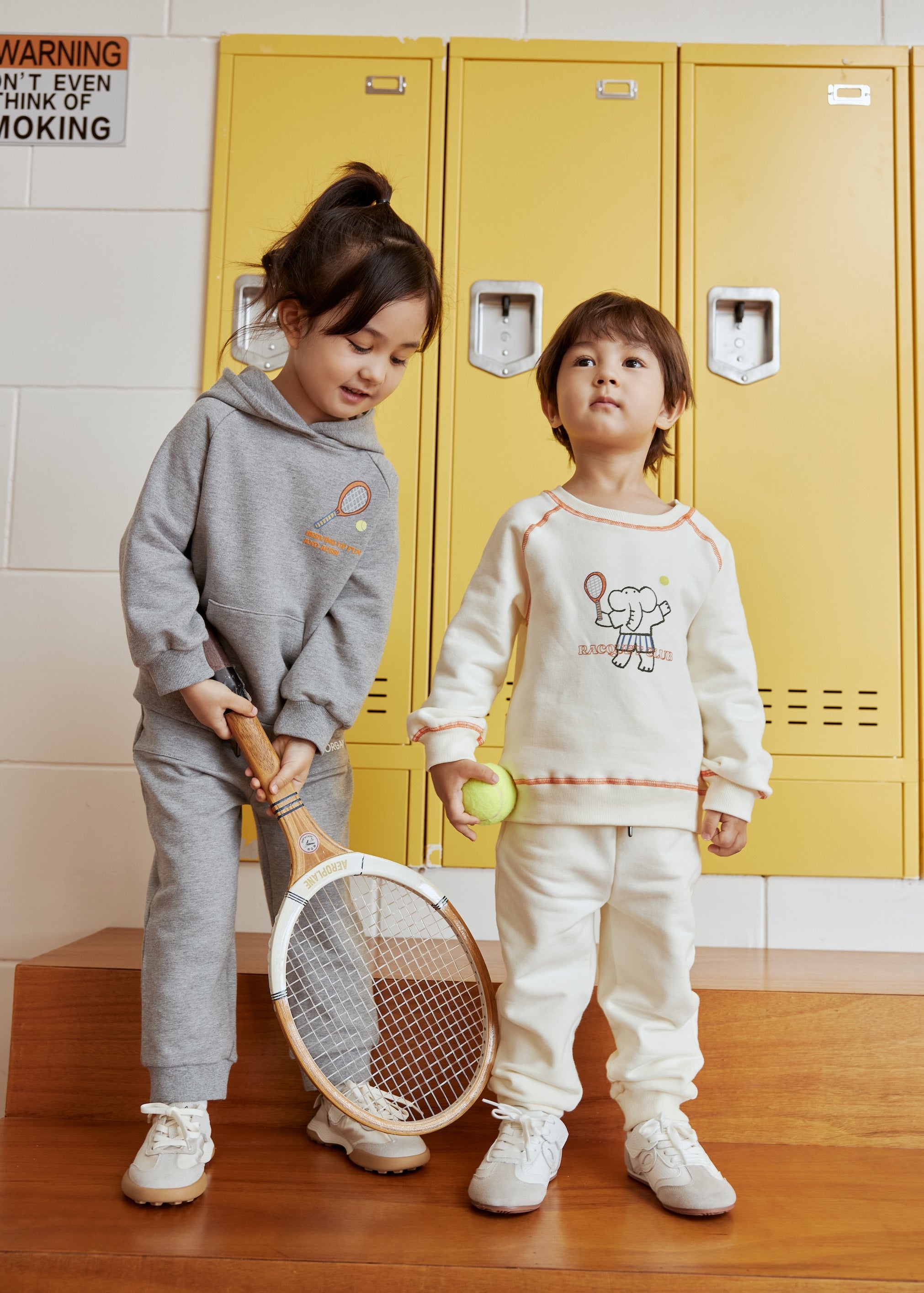 Two children in matching outfits standing in front of yellow lockers.