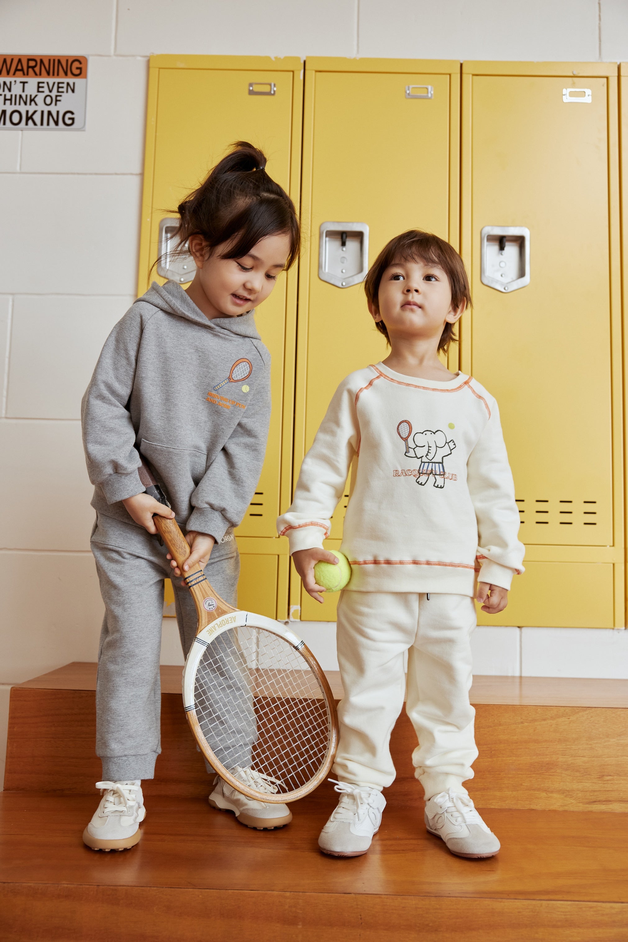 Two children in matching outfits standing in front of yellow lockers.