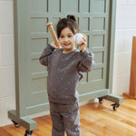 Child holding a baseball and bat in front of a locker with game statistics on a wooden floor.