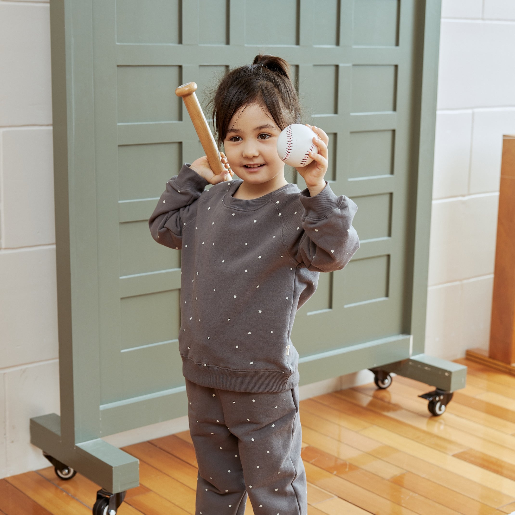 Child holding a baseball and bat in front of a locker with game statistics on a wooden floor.