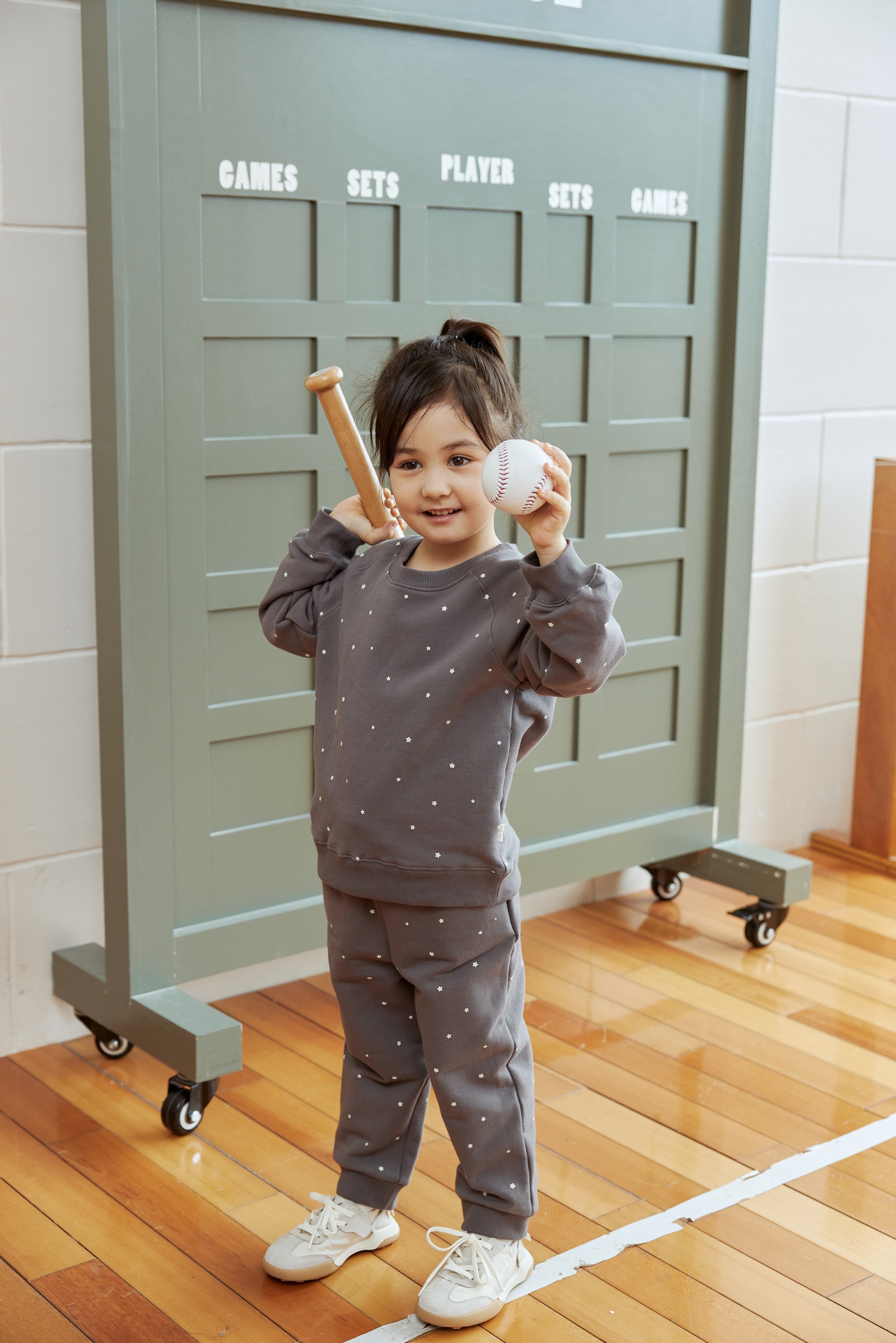Child holding a baseball and bat in front of a locker with game statistics on a wooden floor.