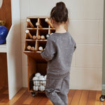 Child in a gray outfit standing next to a wooden toy storage unit with balls on a wooden floor.