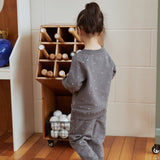 Child in a gray outfit standing next to a wooden toy storage unit with balls on a wooden floor.