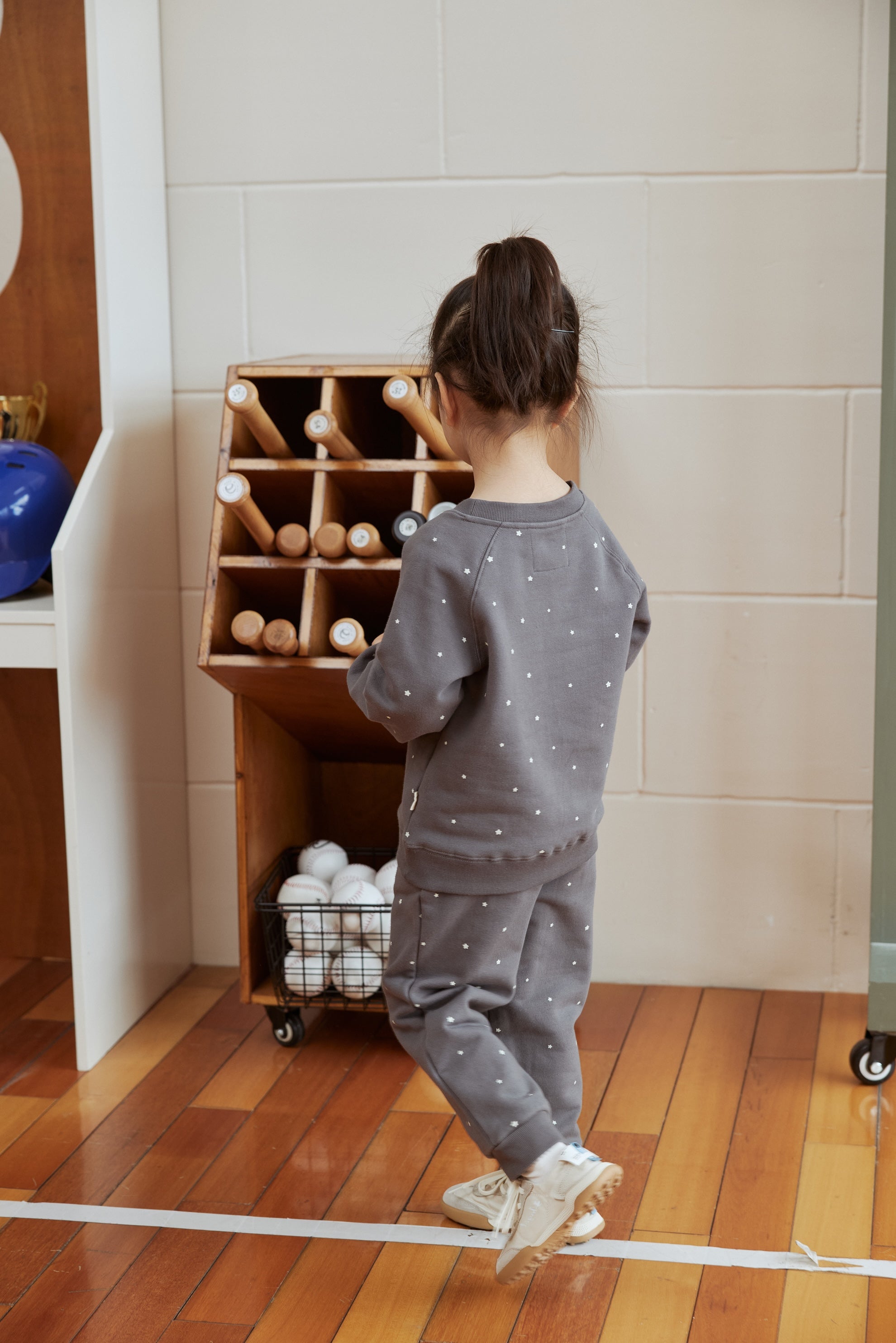 Child in a gray outfit standing next to a wooden toy storage unit with balls on a wooden floor.