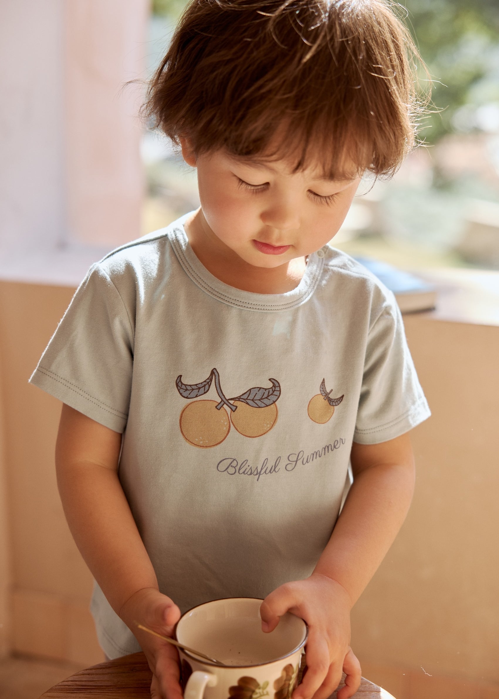a young boy sit beside table with a cup in his hand and wearing Toddler Organic Graphic T-shirt-Clementine