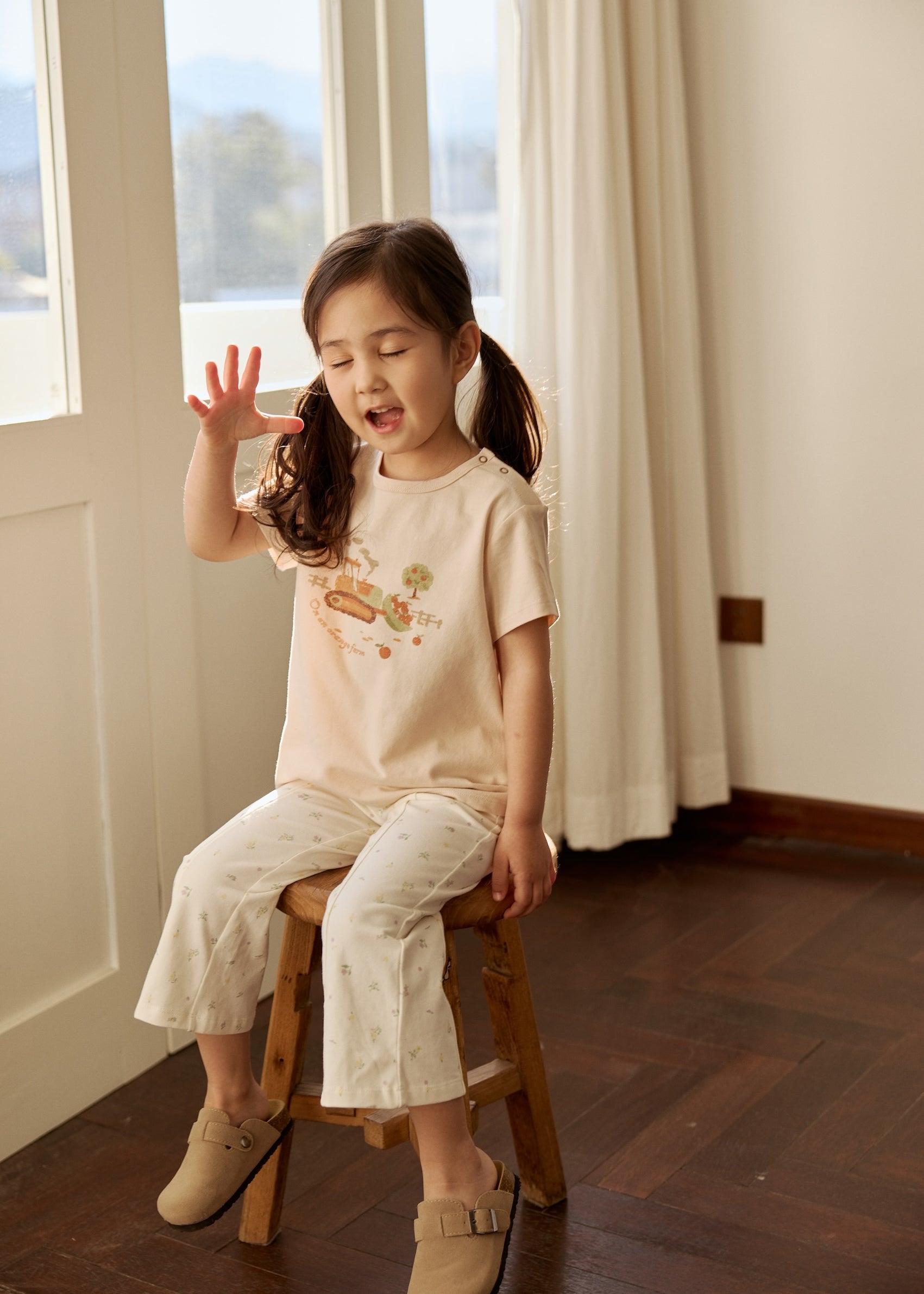 A little girl in a truck graphic tee sits on a chair, raising her hand with a cheerful expression.
