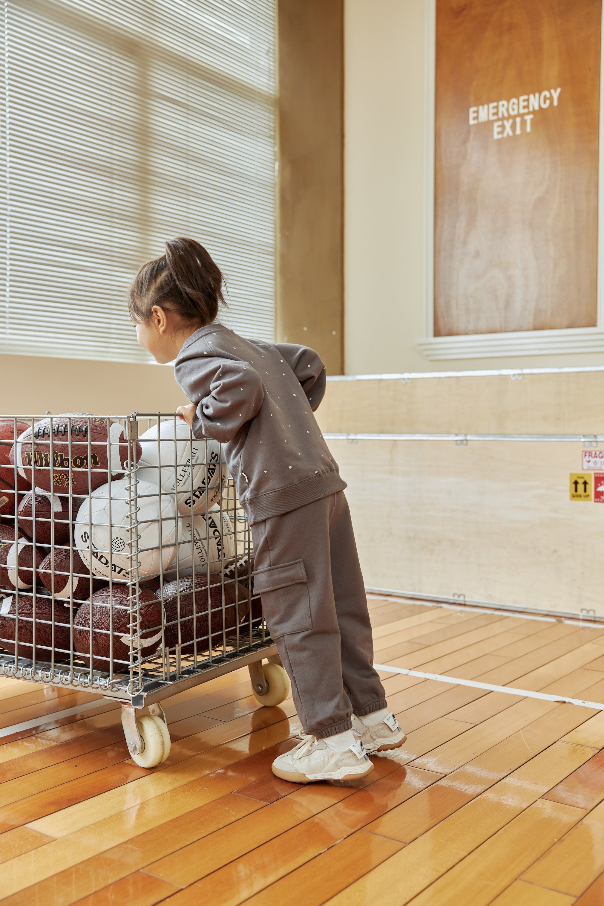 Child pushing a cart full of basketballs in an indoor setting with an emergency exit sign visible.