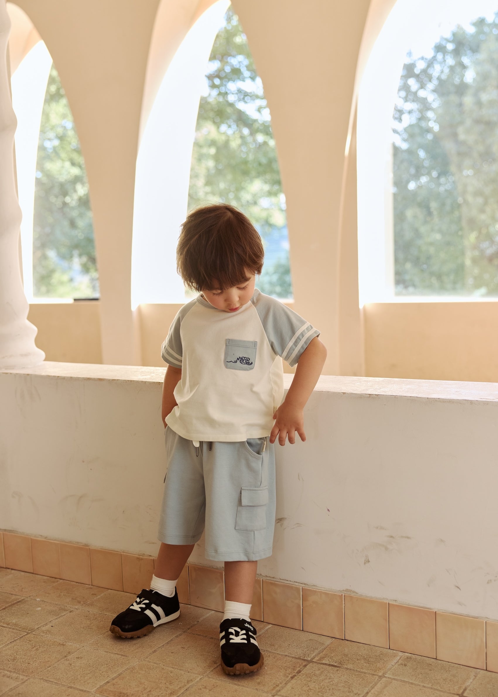 A young boy in toddler Cargo Shorts-Light Blue stands by a sidewall, looking down to his feet