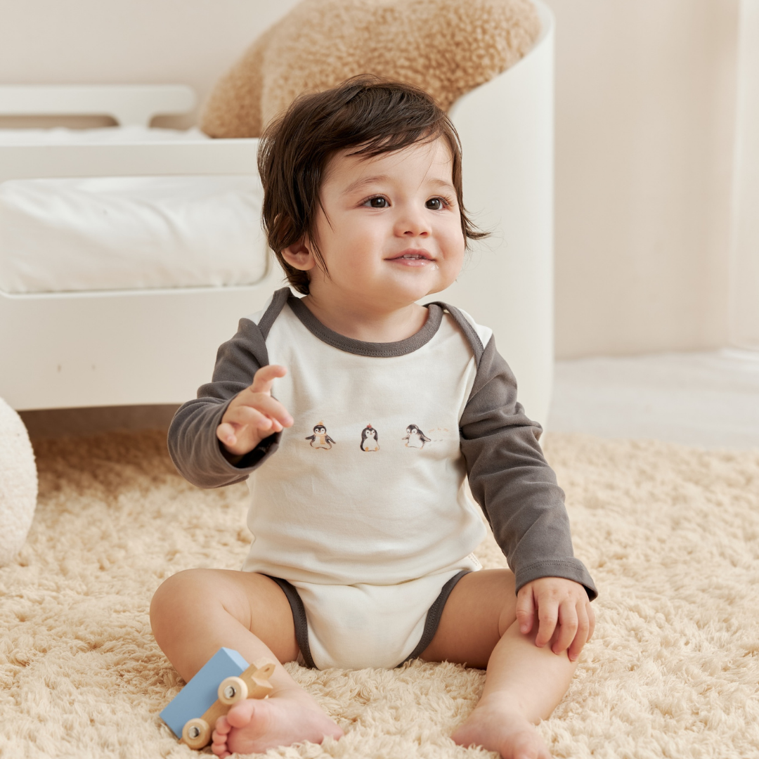 Baby sitting on a carpeted floor wearing a onesie with penguin designs.
