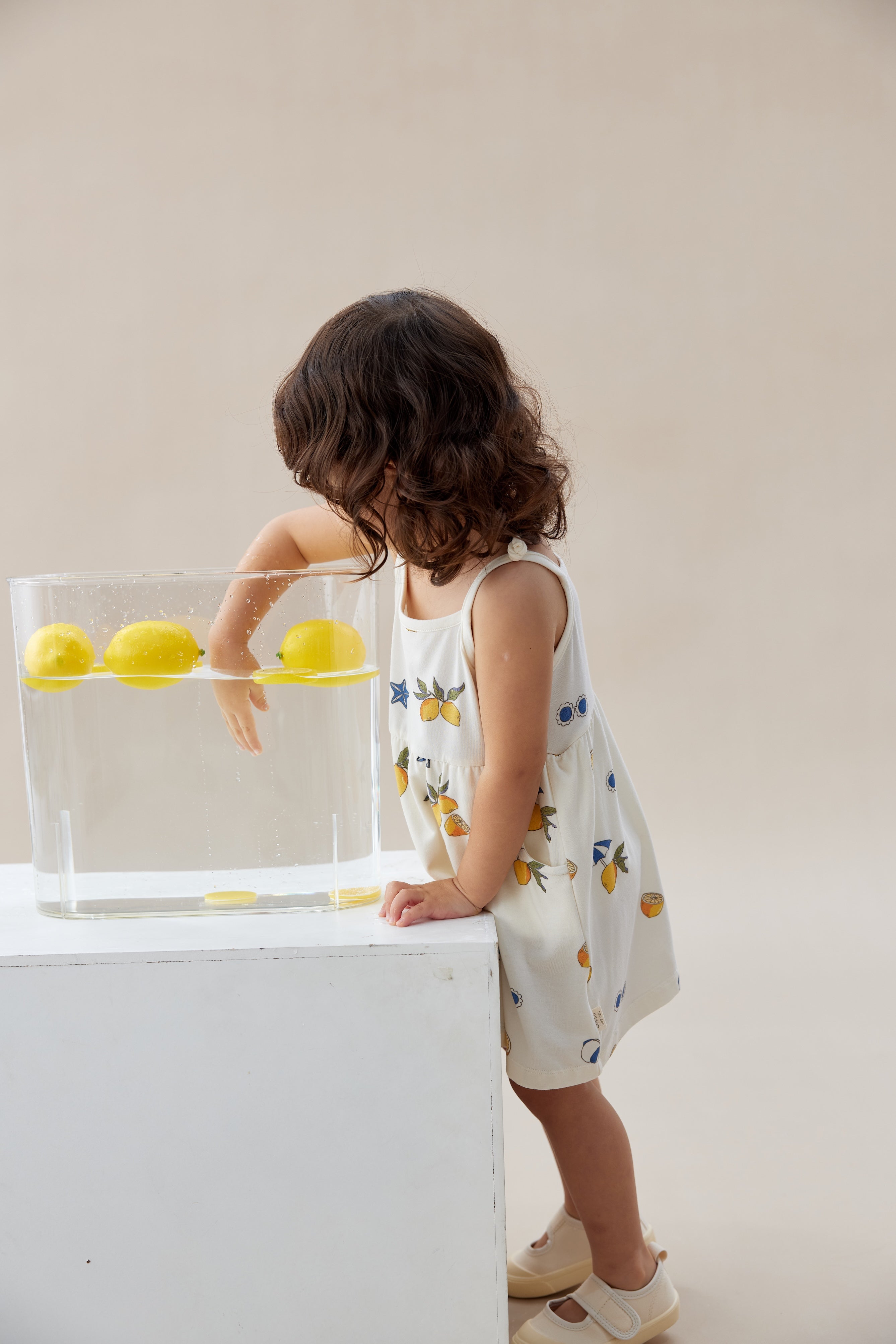 Child in a sleeveless dress with lemon pattern interacting with a transparent box on a white surface.