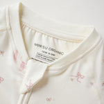 Close-up of a baby romper or long-sleeve top in warm off-white/cream fabric with small pink bow prints and a visible inner neck label reading 'Norsu organic