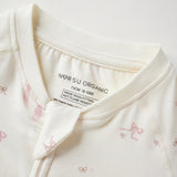 Close-up of a baby romper or long-sleeve top in warm off-white/cream fabric with small pink bow prints and a visible inner neck label reading 'Norsu organic