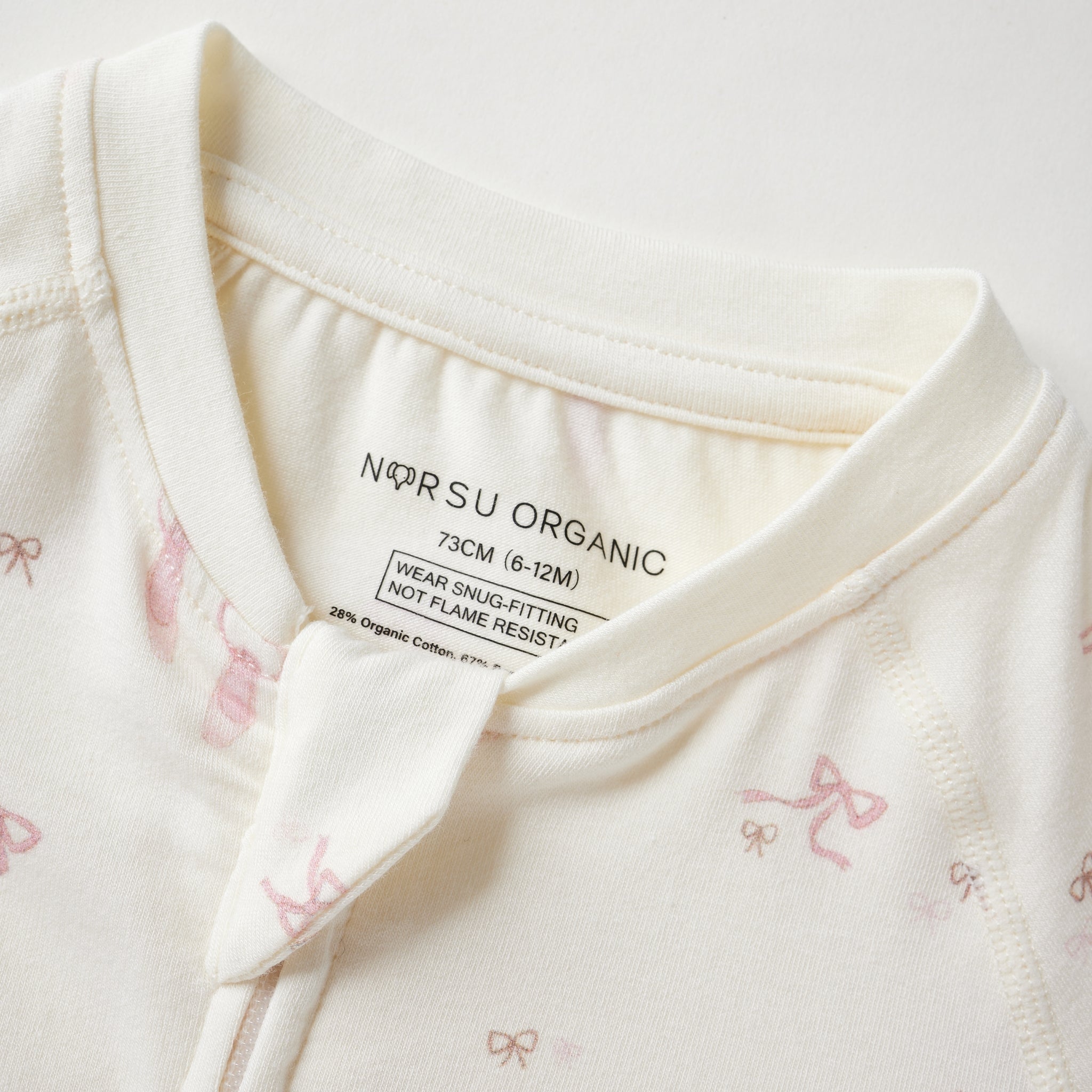 Close-up of a baby romper or long-sleeve top in warm off-white/cream fabric with small pink bow prints and a visible inner neck label reading 'Norsu organic
