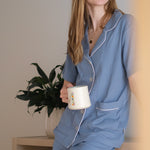 Woman in blue pajamas holding a mug, sitting on a wooden stool with a plant in the background.