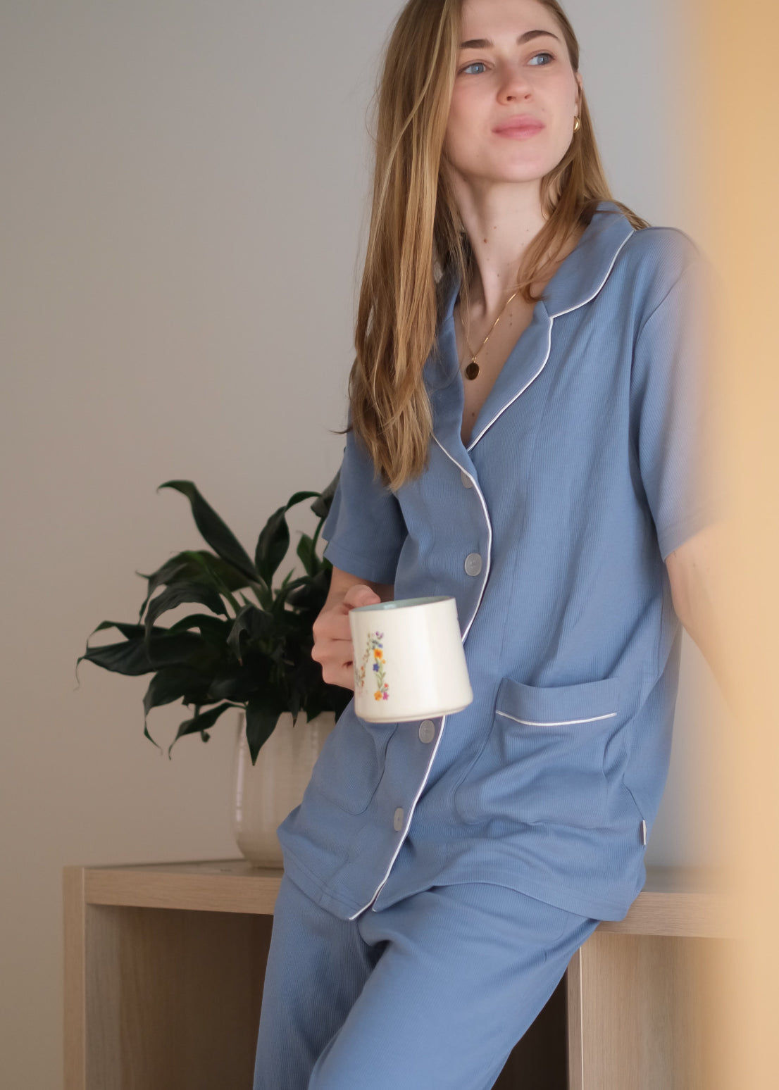 Woman in blue pajamas holding a mug, sitting on a wooden stool with a plant in the background.