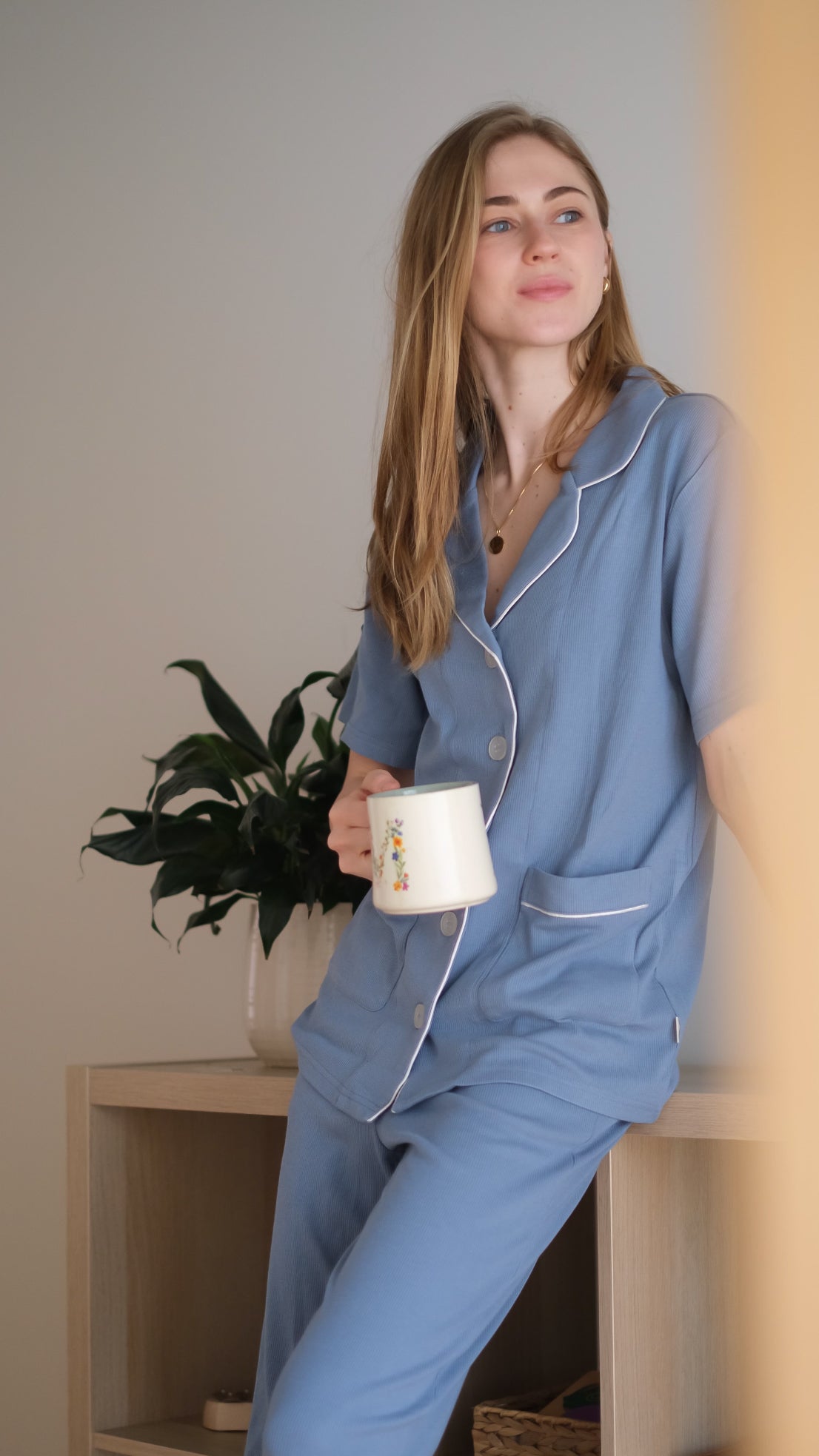 Woman in blue pajamas holding a mug, sitting on a wooden stool with a plant in the background.