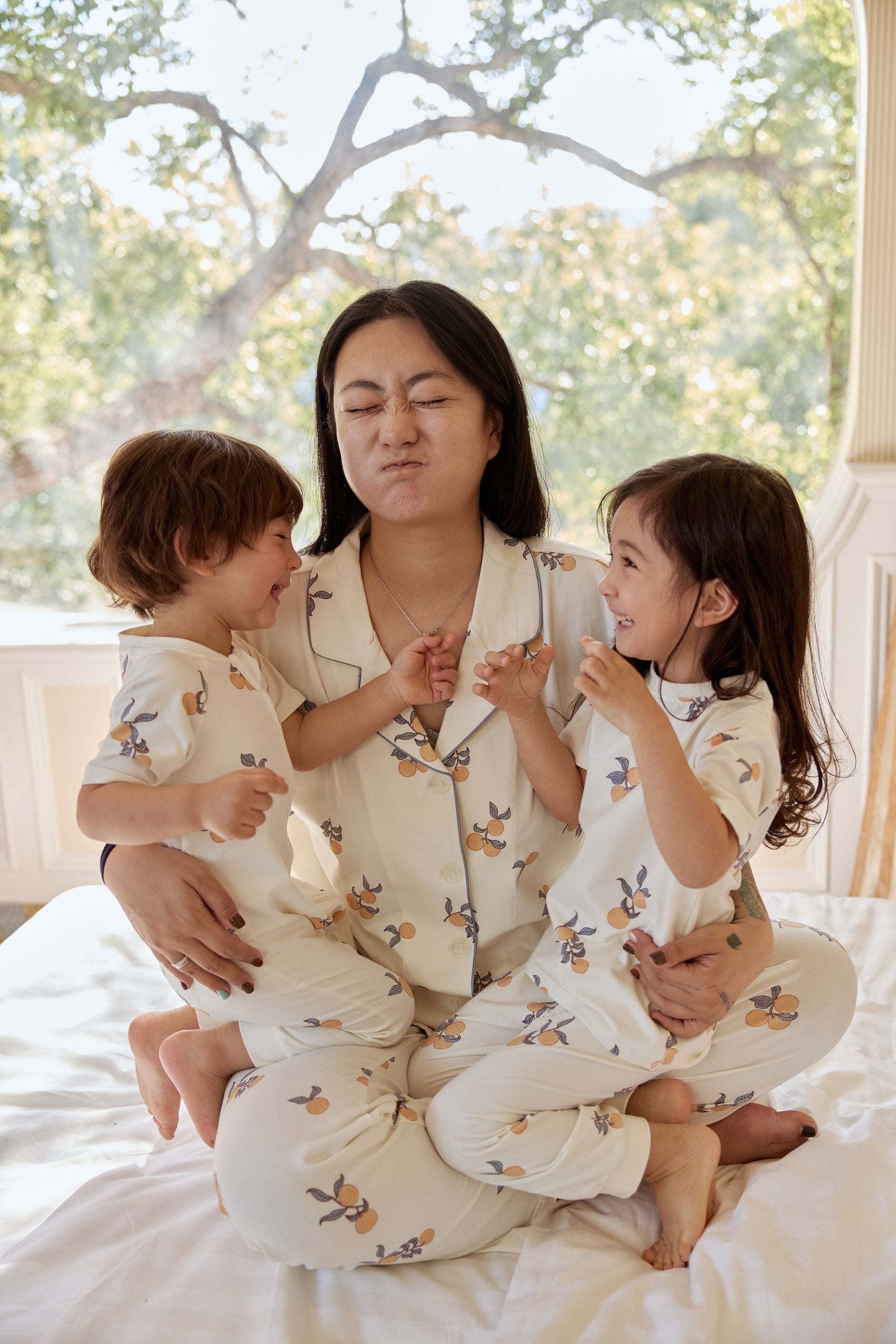 A woman and her two kids sit happily on a bed, all wearing soft summer pajamas, creating a warm family moment