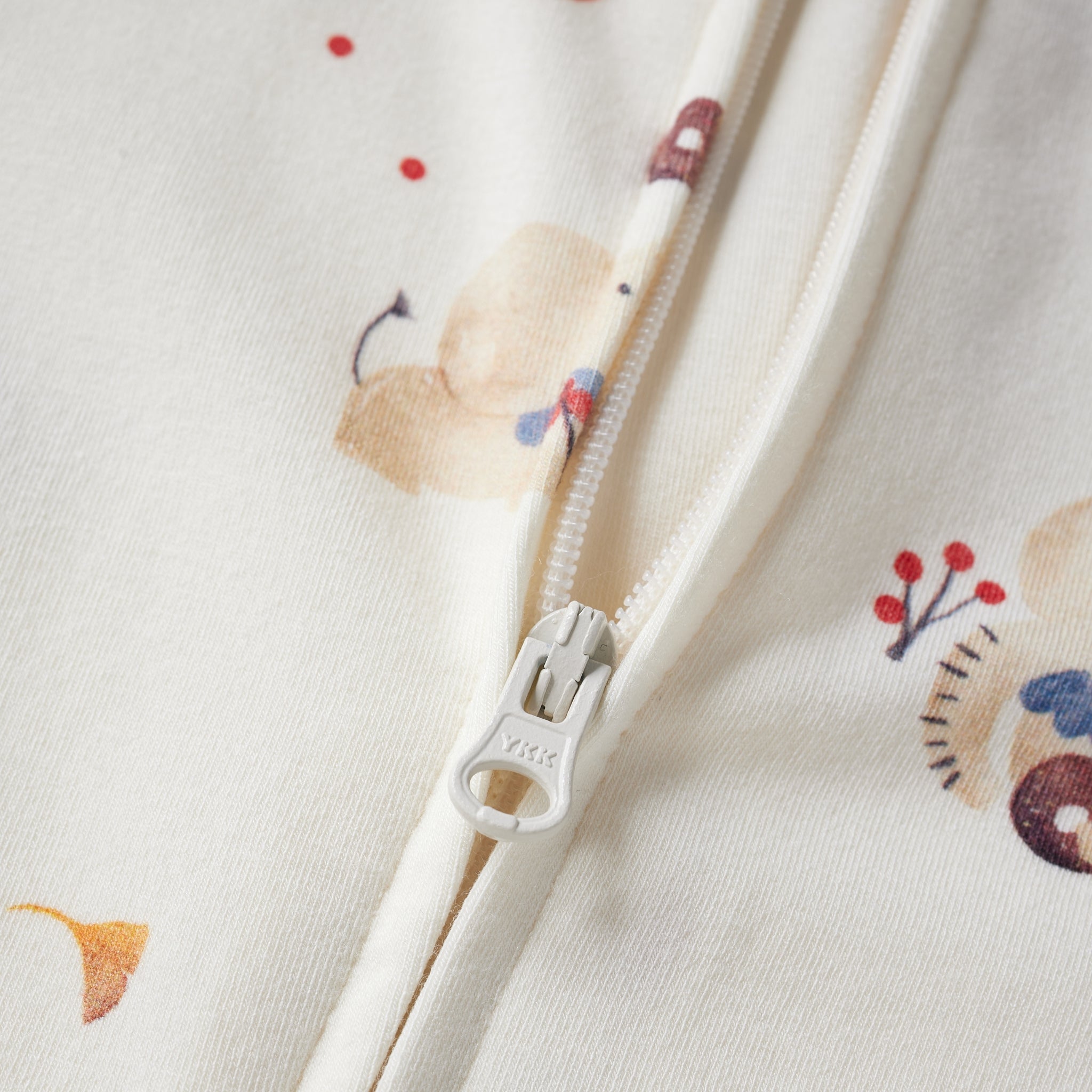 Close-up of a cream-colored zippered garment with a soft pastel animal and berry print, featuring a white zipper pull with 'YKK' branding.
